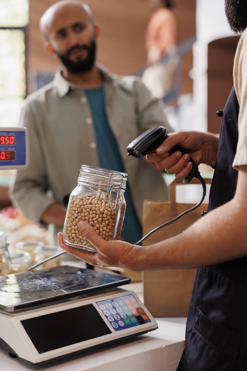 Shopkeeper scanning a packaged item at a counter beside a weighing scale