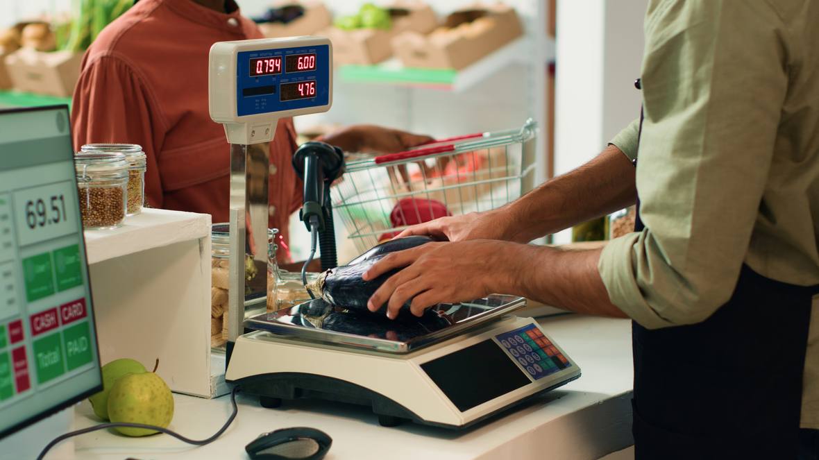 Weighing scale on a shop counter showing weight and unit price beside a point of sale screen